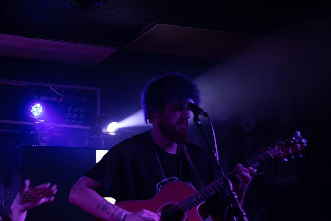 A musician plays an acoustic guitar and sings into a microphone on a dimly lit stage bathed in blue and purple light.