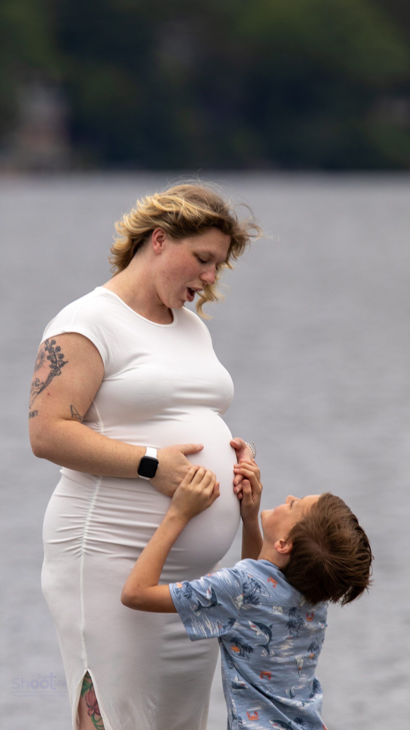 A person wearing a white dress stands by a body of water while a child reaches up to touch their pregnant stomach.