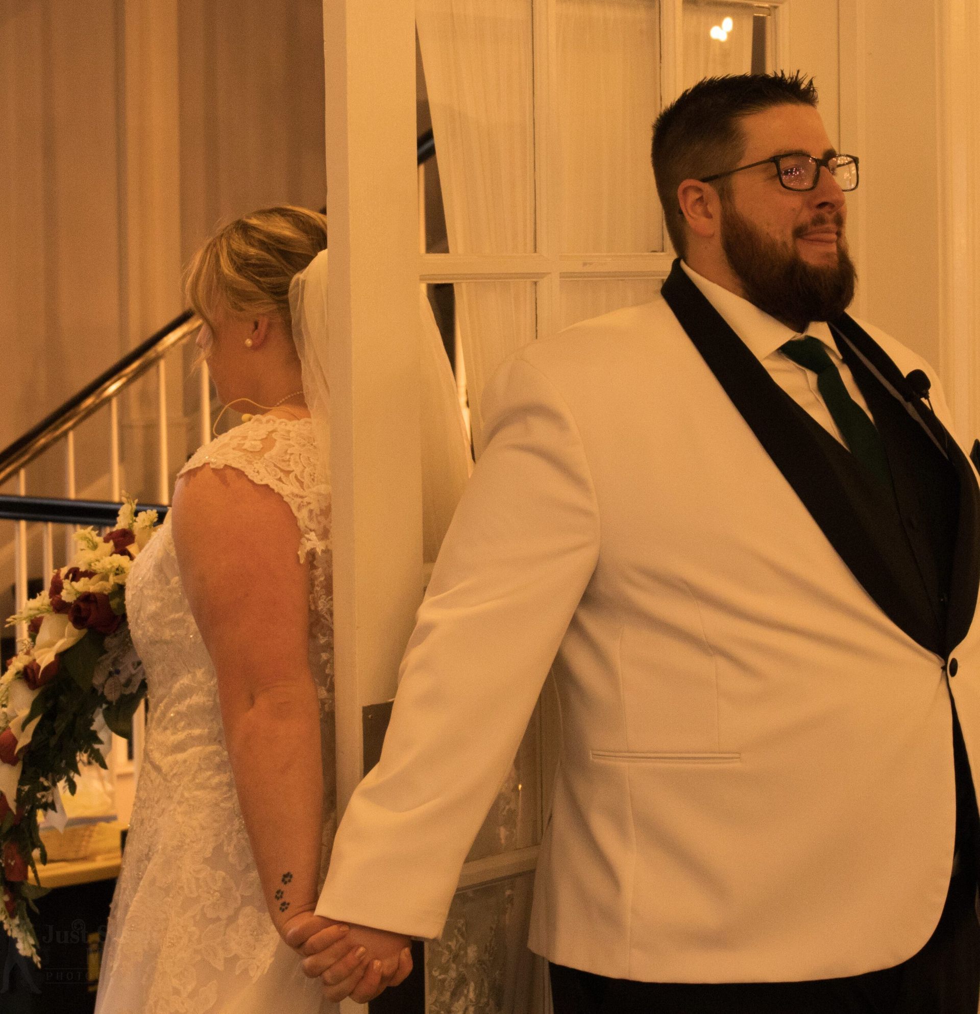 A bride and groom hold hands while separated by a door frame, their faces turned away from each other.