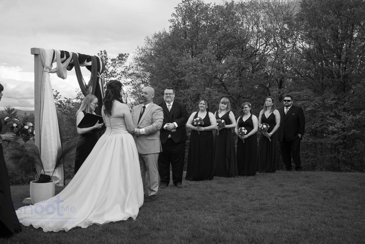 A wedding ceremony outdoors with a couple holding hands before an officiant and a bridal party lined up in a grassy field.