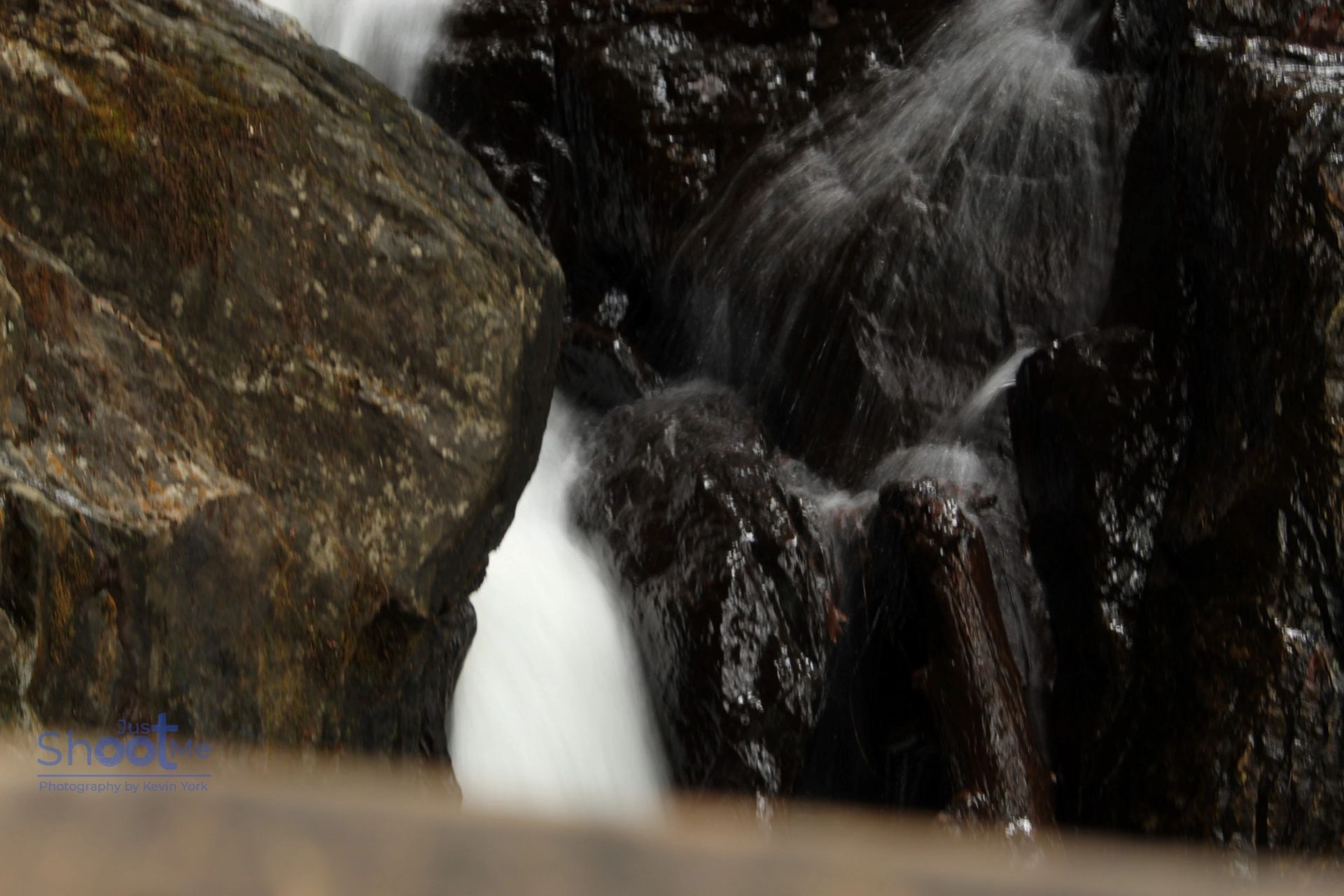 Water cascades through a narrow gap between dark, wet rocks in a rocky stream.