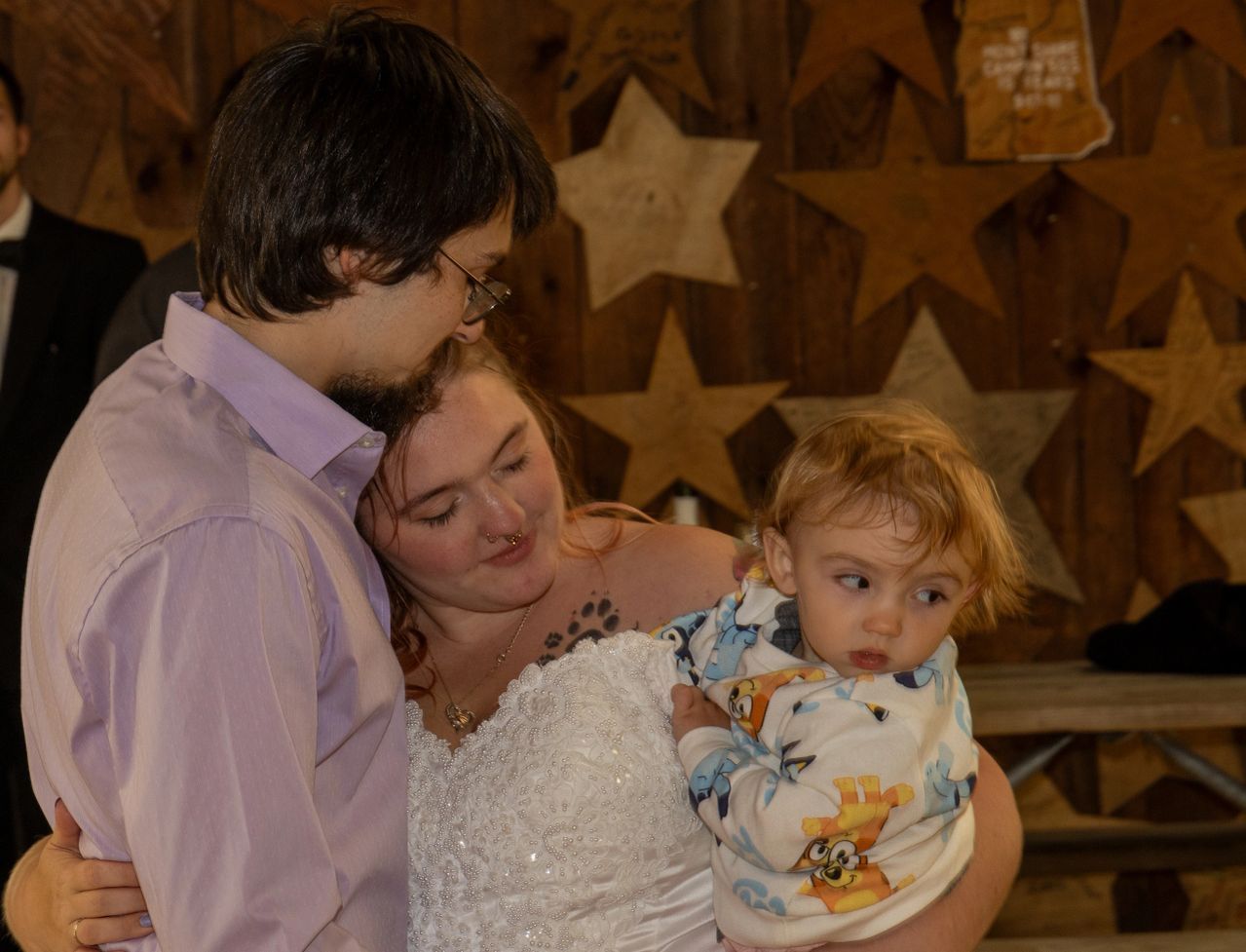 A person in a light purple shirt and a person in a white wedding dress hold a toddler in front of a wall of wooden stars.