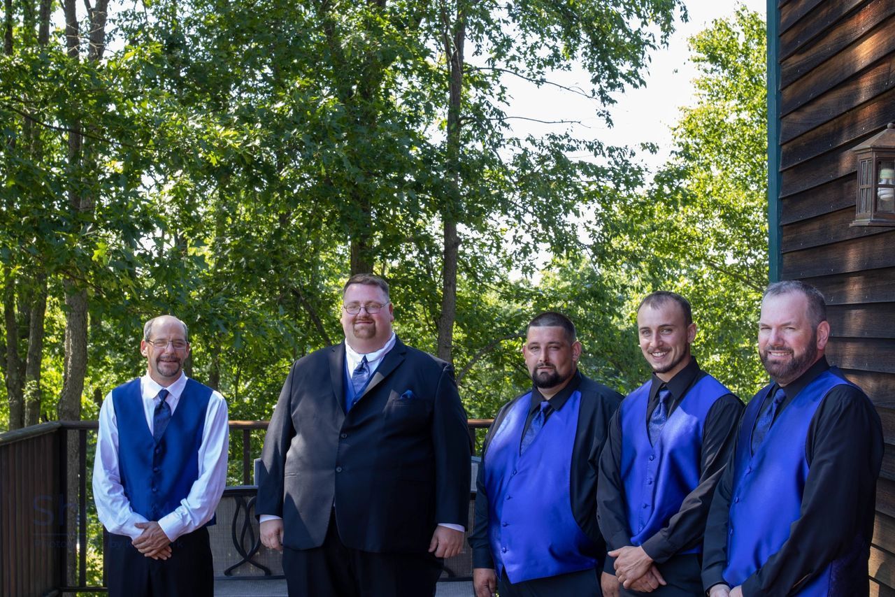 Five men in blue vests and ties pose for a formal portrait outdoors on a wooden deck with trees in the background.