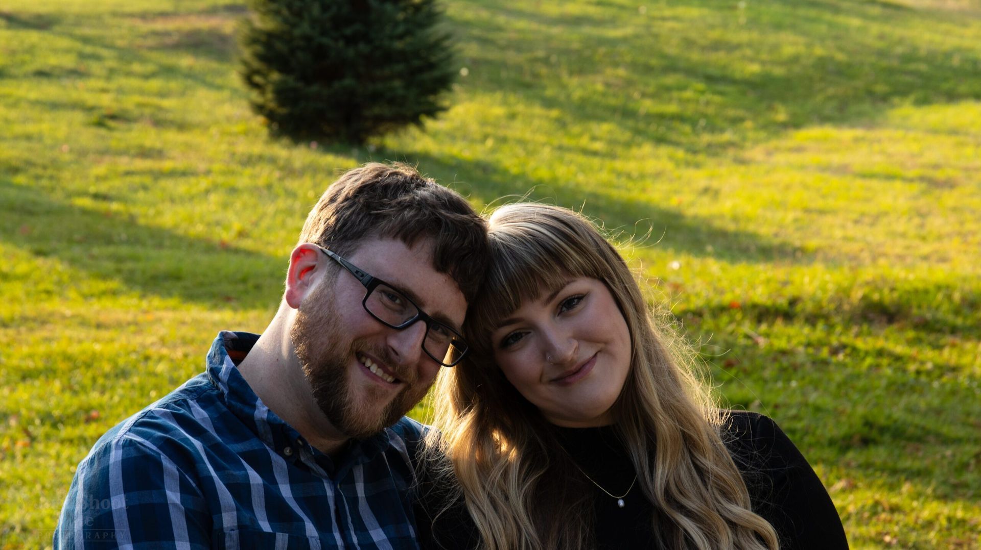 A person with glasses and a plaid shirt leans their head against a smiling person with long hair in a grassy outdoor field.