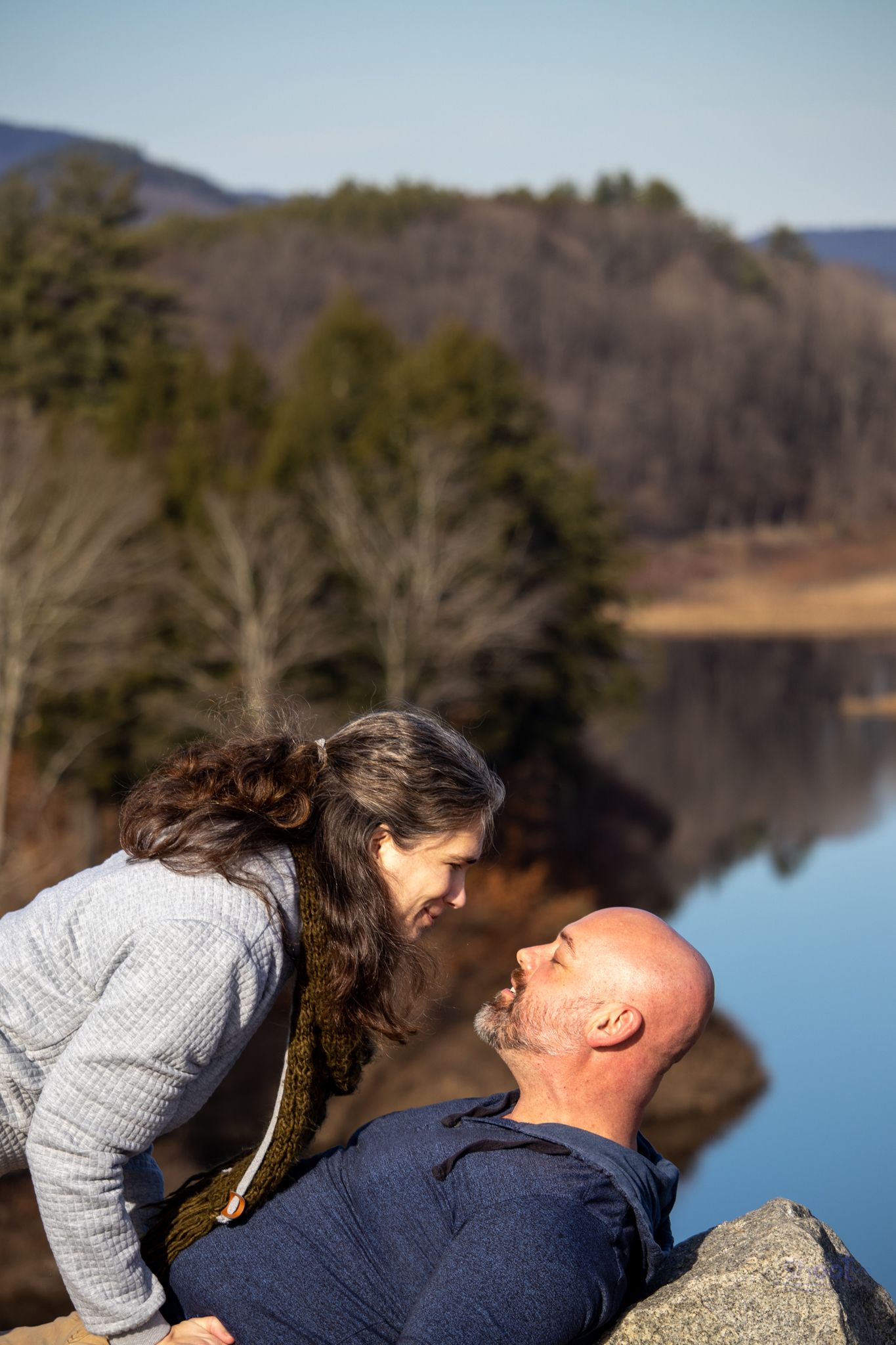 A person looking down at another leaning back against a rock, with a calm lake and treed mountains in the background.