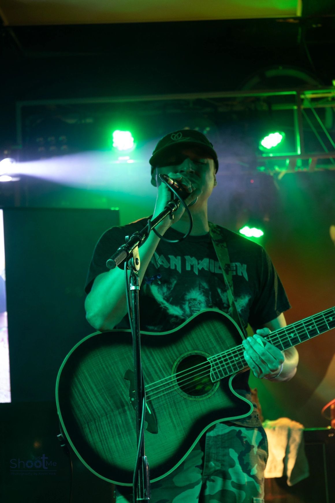 A musician in a cap and band t-shirt performs on stage with an acoustic guitar, illuminated by bright green stage lights.