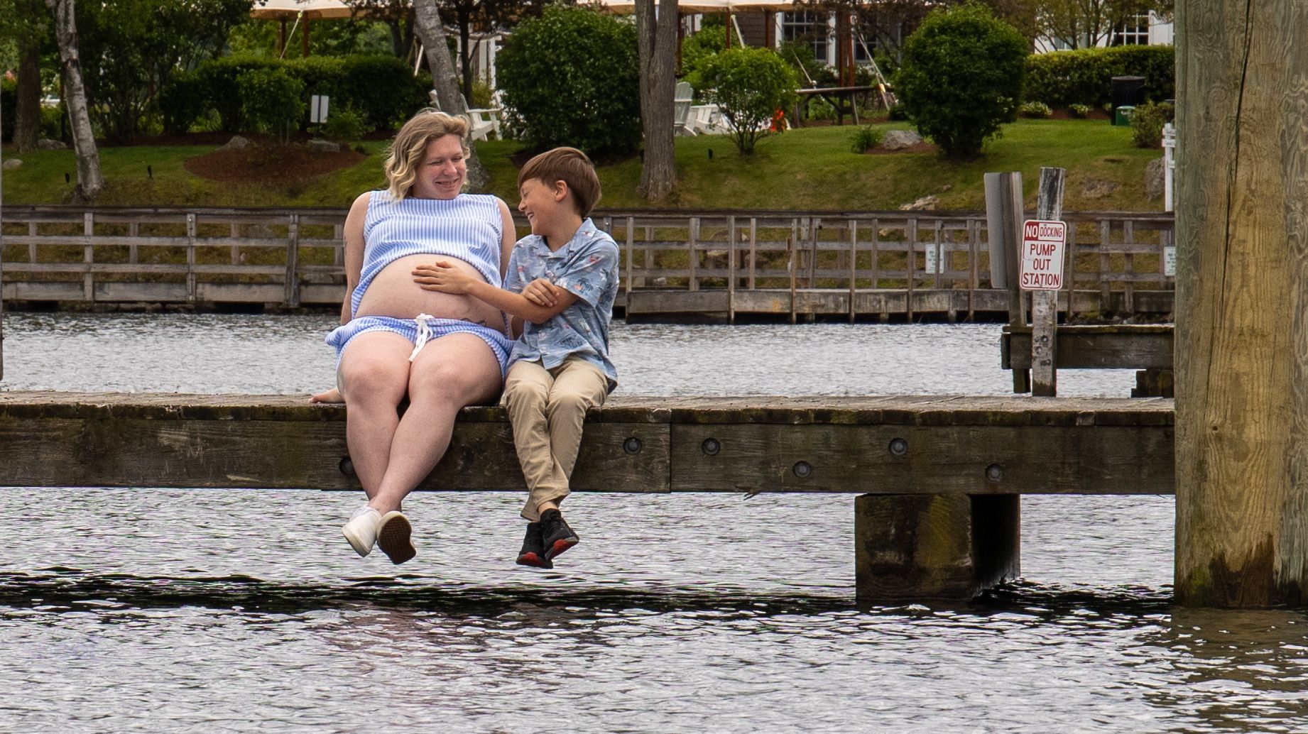 A pregnant woman and a young boy sit together on a wooden pier by the water, looking at each other and smiling.