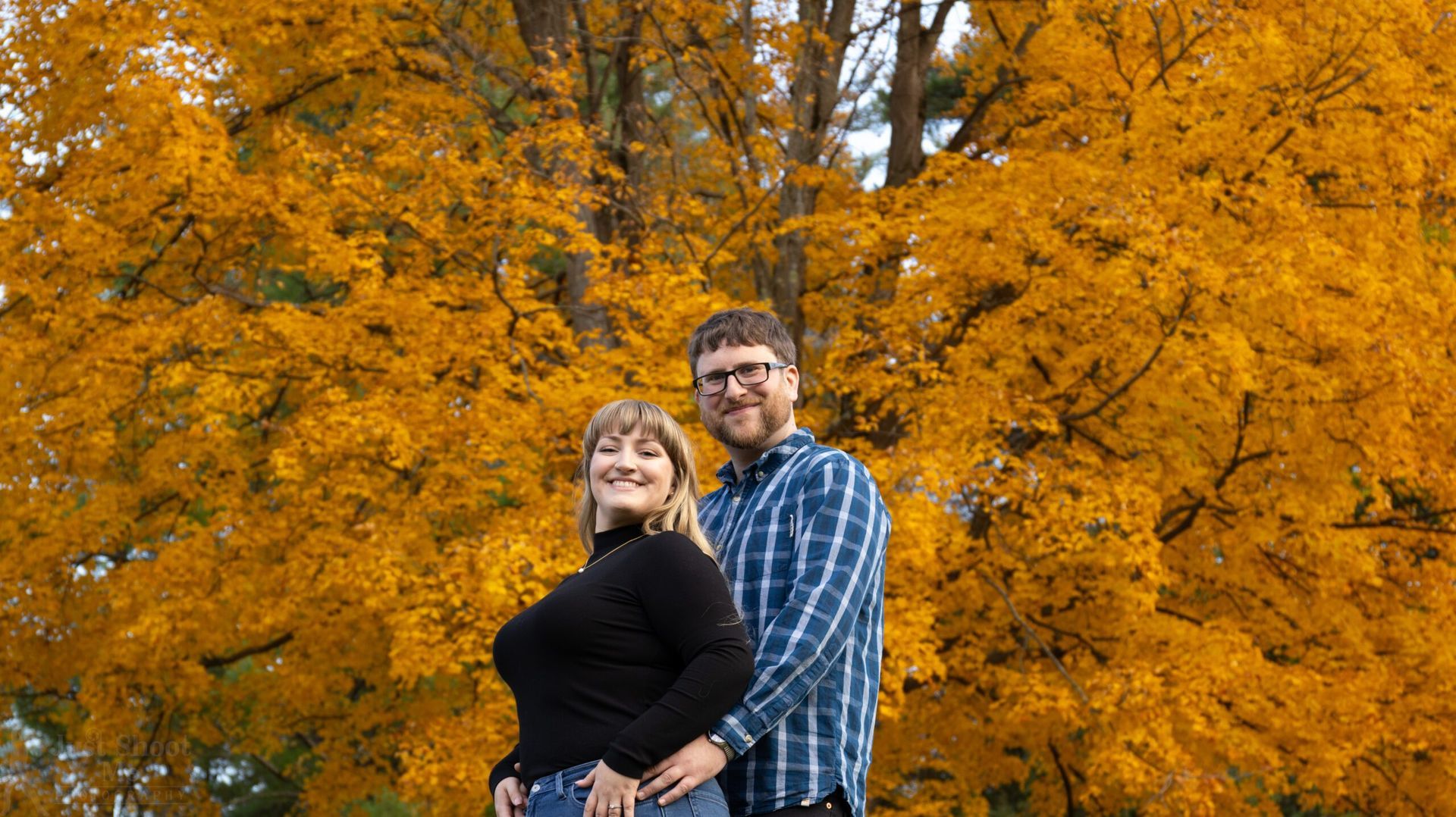 A couple standing together in front of a tree with vibrant yellow autumn leaves.