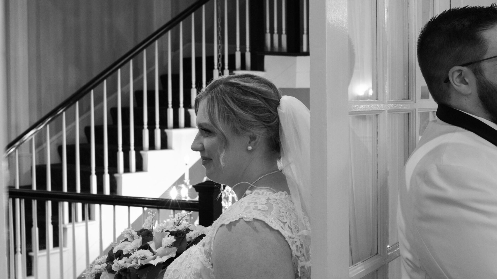 A bride and groom stand back-to-back near a glass door and staircase, participating in a pre-ceremony wedding moment.