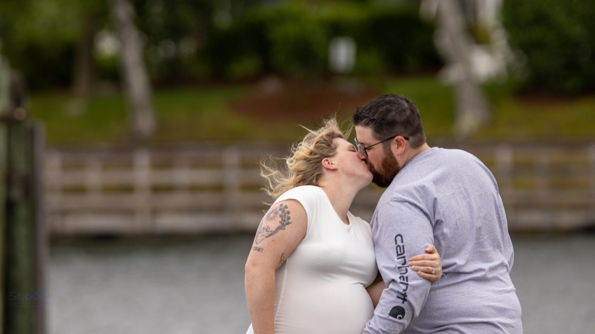 A couple kisses while standing outdoors near a body of water with a wooden fence in the background.