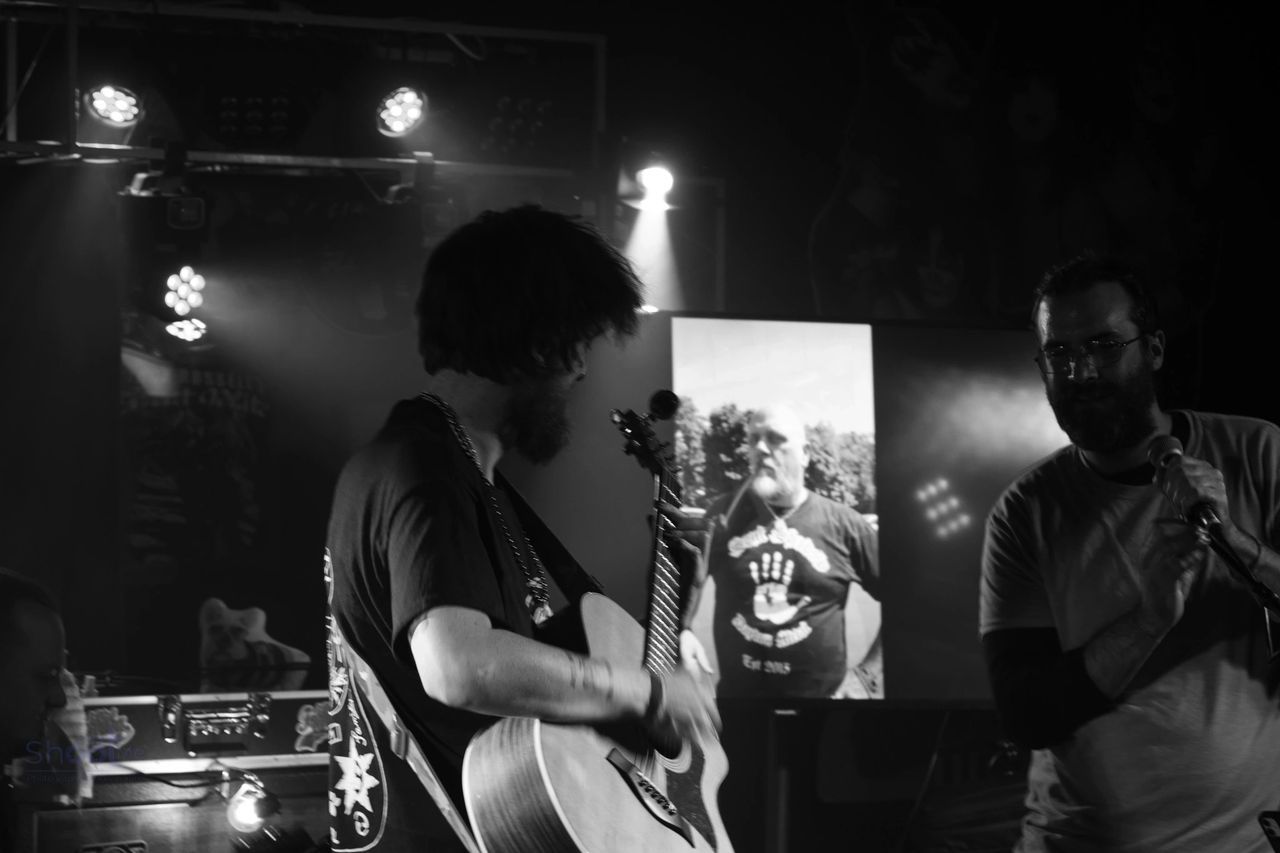 A performer plays an acoustic guitar on a stage with a screen displaying a photo of a person in the background.