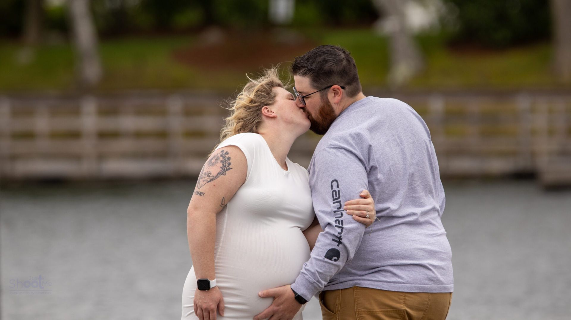 A pregnant person and their partner kiss near a lake, standing on a wooden walkway during an outdoor photo session.
