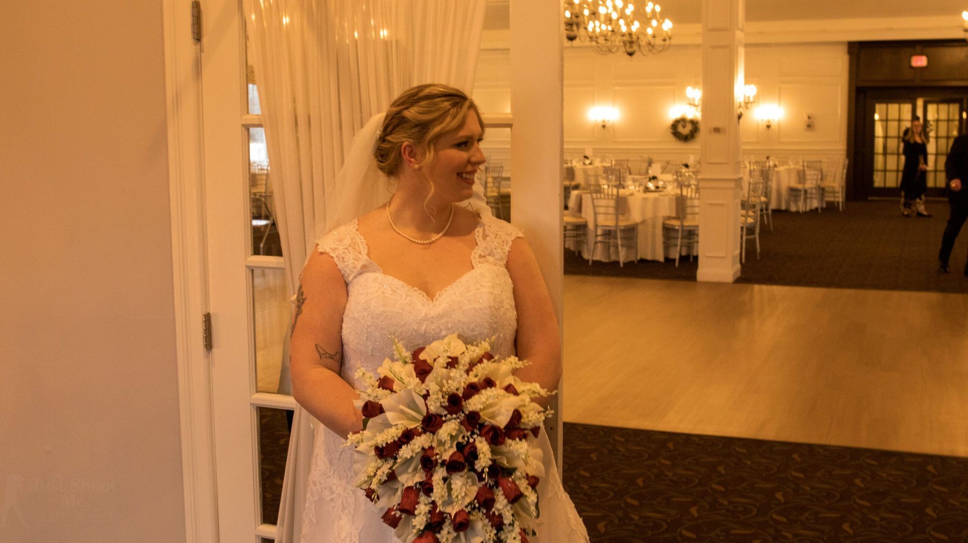 A bride in a white wedding dress holds a bouquet of white and red roses while smiling inside a formal event hall.