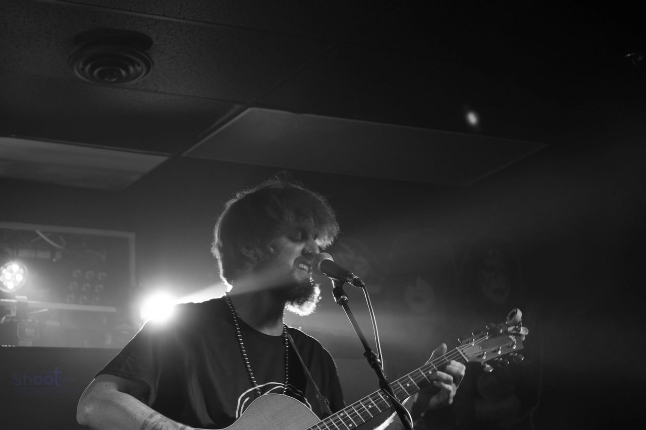 A musician singing into a microphone while playing an acoustic guitar on a dimly lit, stage-like setting.