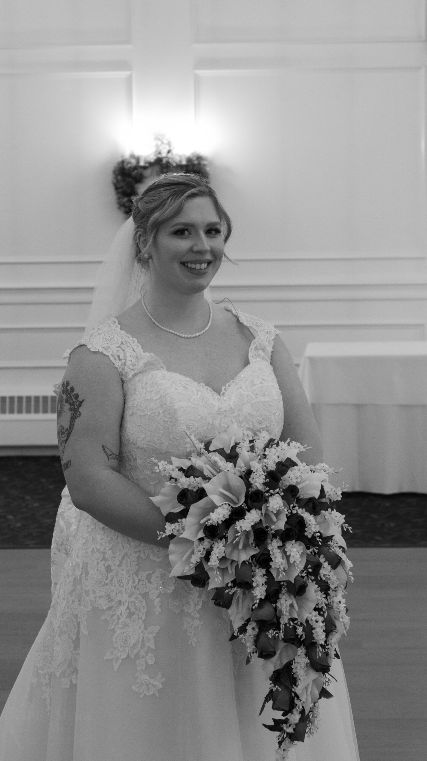 A person in a lace wedding gown holding a large cascading bouquet, smiling in front of a white paneled wall.
