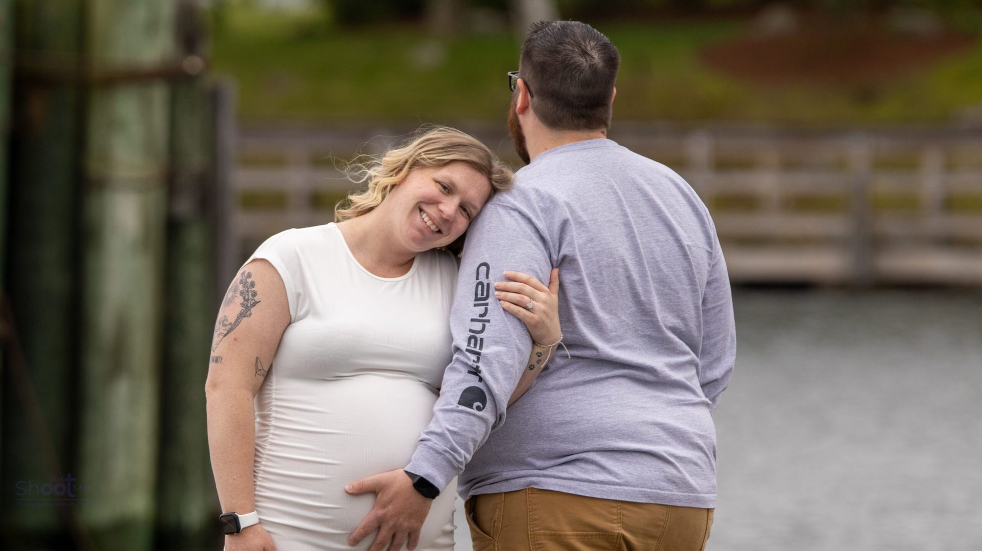 A couple embraces outdoors near a pier, with the smiling partner leaning on the other's shoulder.