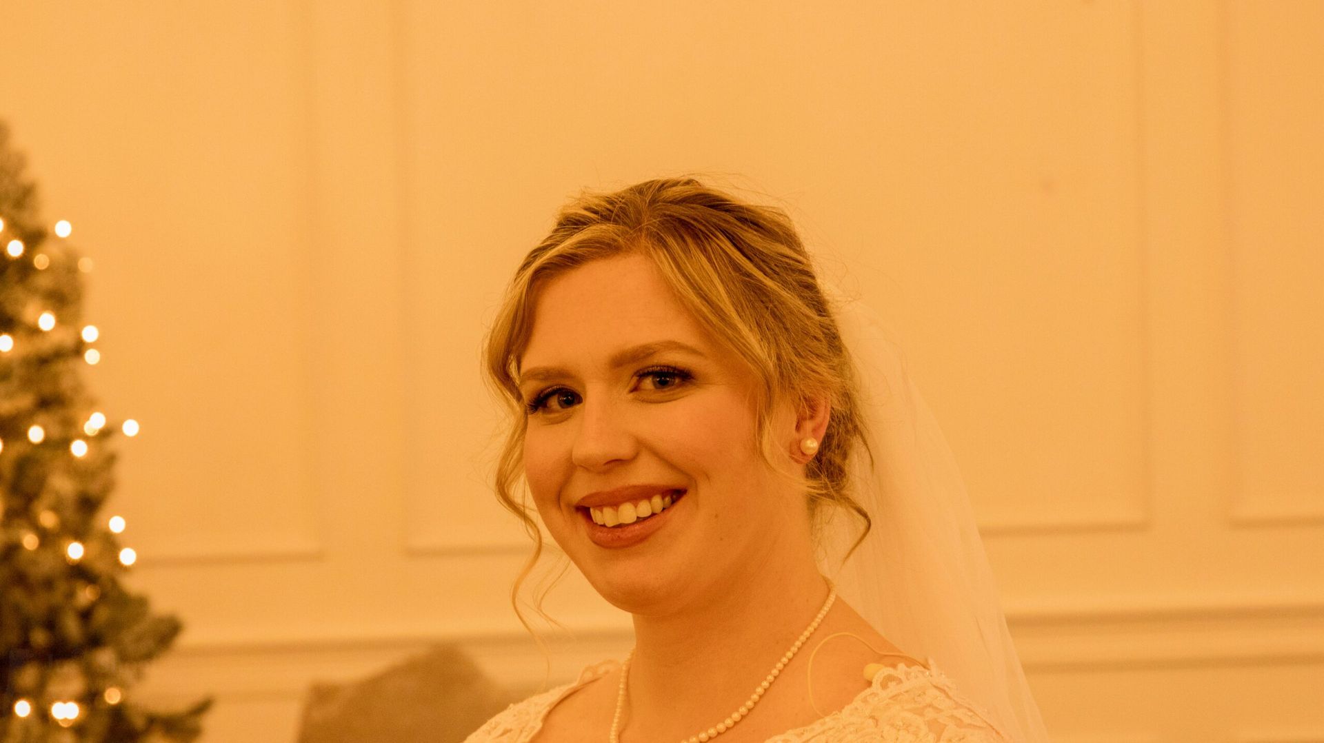 A smiling person wearing a white bridal dress and veil, posed in front of a softly lit Christmas tree.