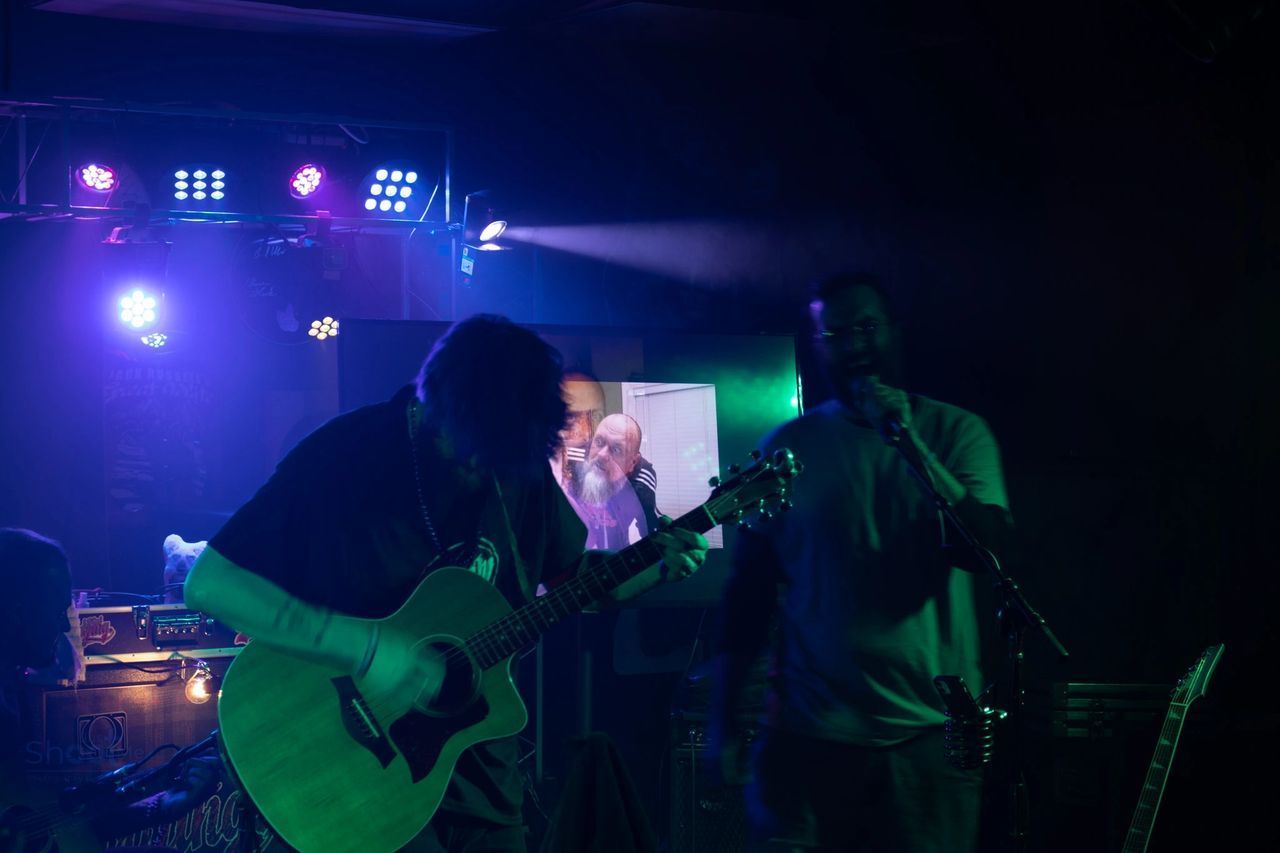 A musician plays an acoustic guitar on a dimly lit stage with blue stage lighting, joined by a second person nearby.