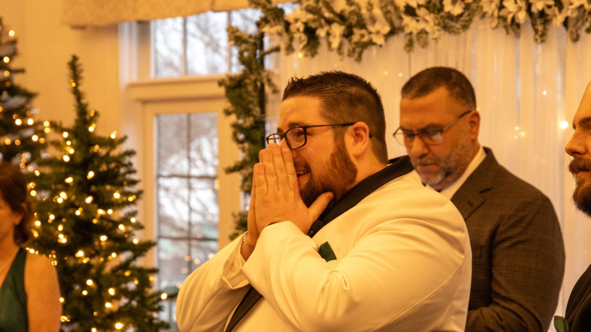 A man in a white suit with glasses reacts with emotion, hands clasped near his face, standing before a lit holiday tree.