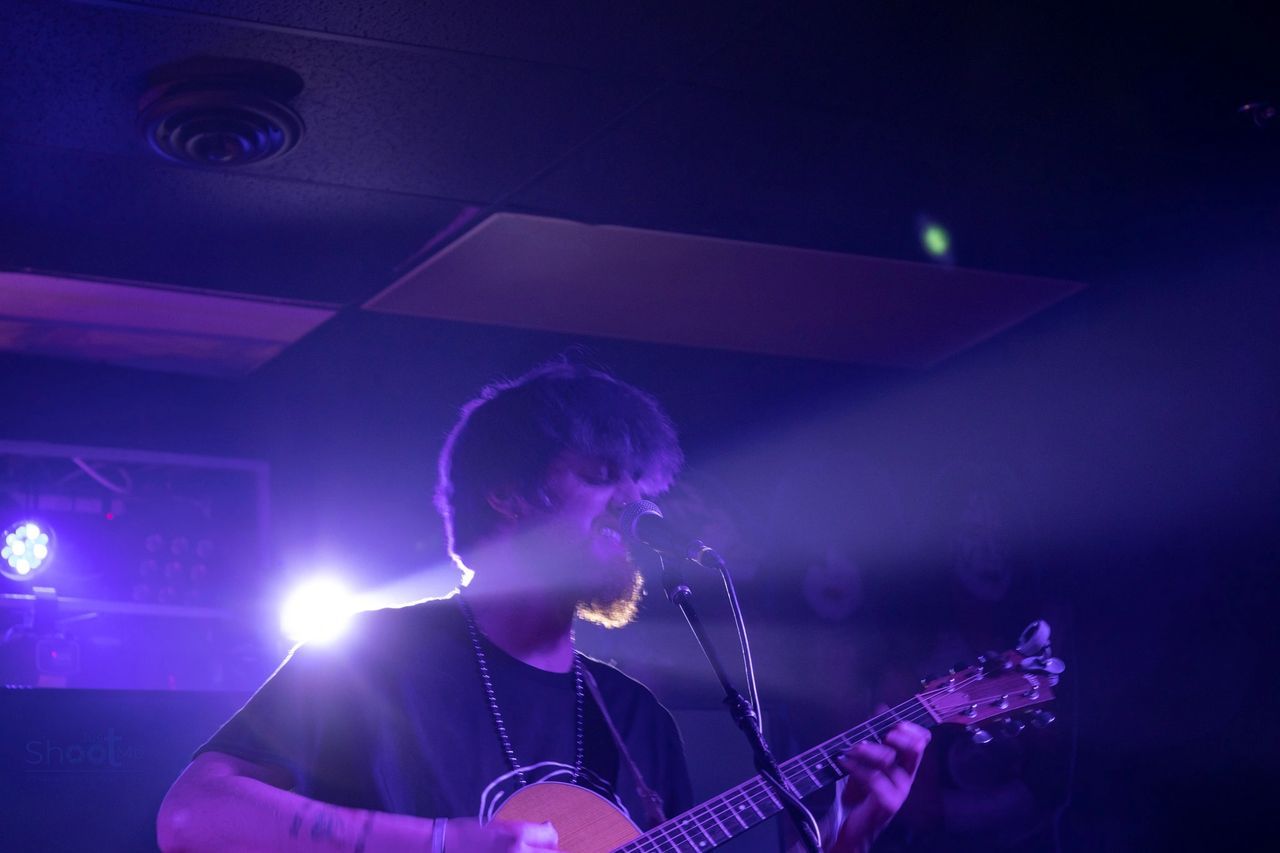 A musician with messy hair plays an acoustic guitar while singing into a microphone under dramatic purple stage lights.