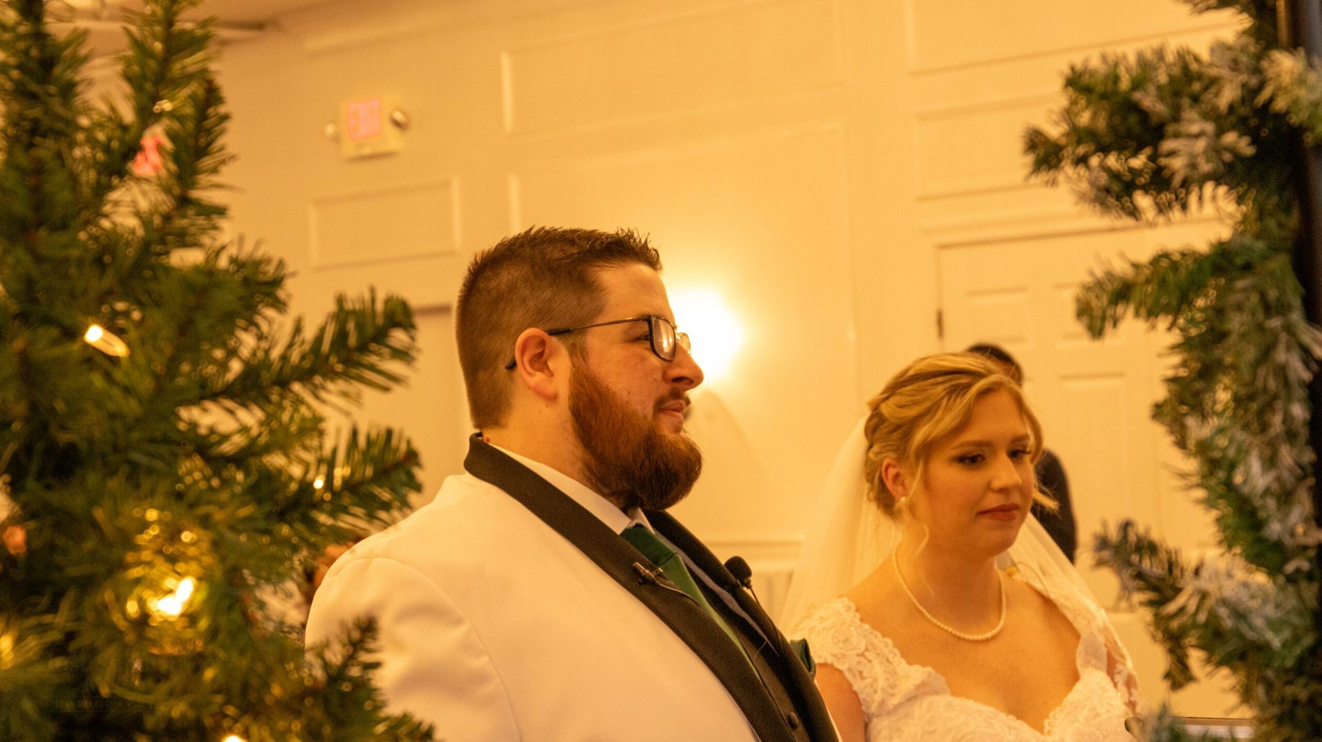 A bride and groom stand together in wedding attire, framed by decorated greenery in a warmly lit indoor setting.