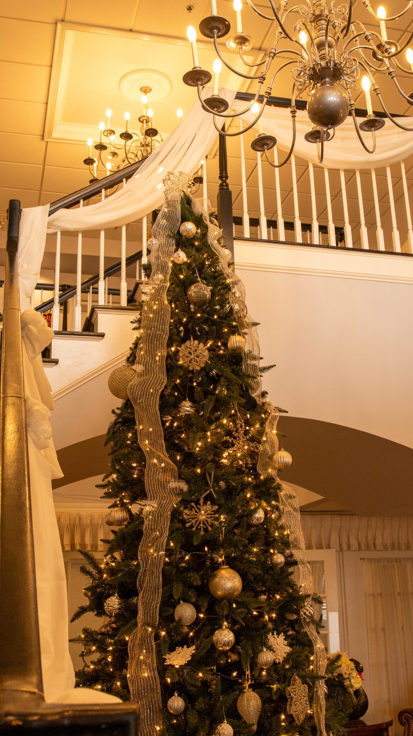 A tall, decorated Christmas tree stands at the base of a grand staircase, accented with gold ribbons and warm lights.