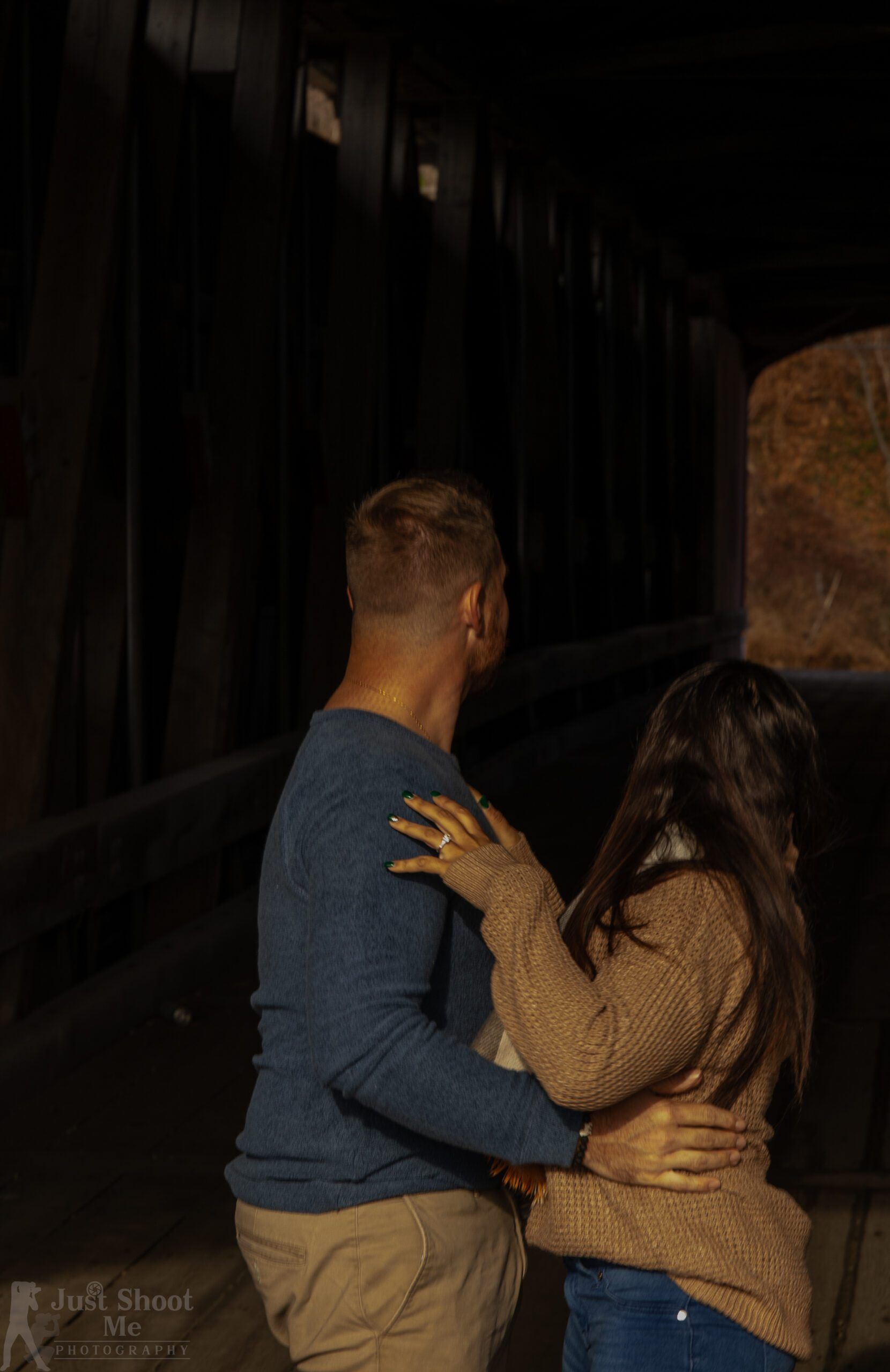 A couple stands close together in a dark, wooden-slatted covered bridge, looking toward the light at the tunnel exit.