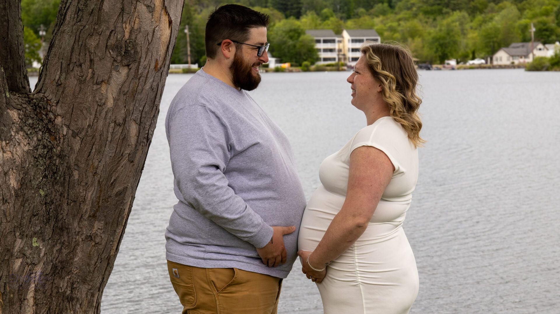 A couple standing by a lake, both holding their bellies, smiling at each other.