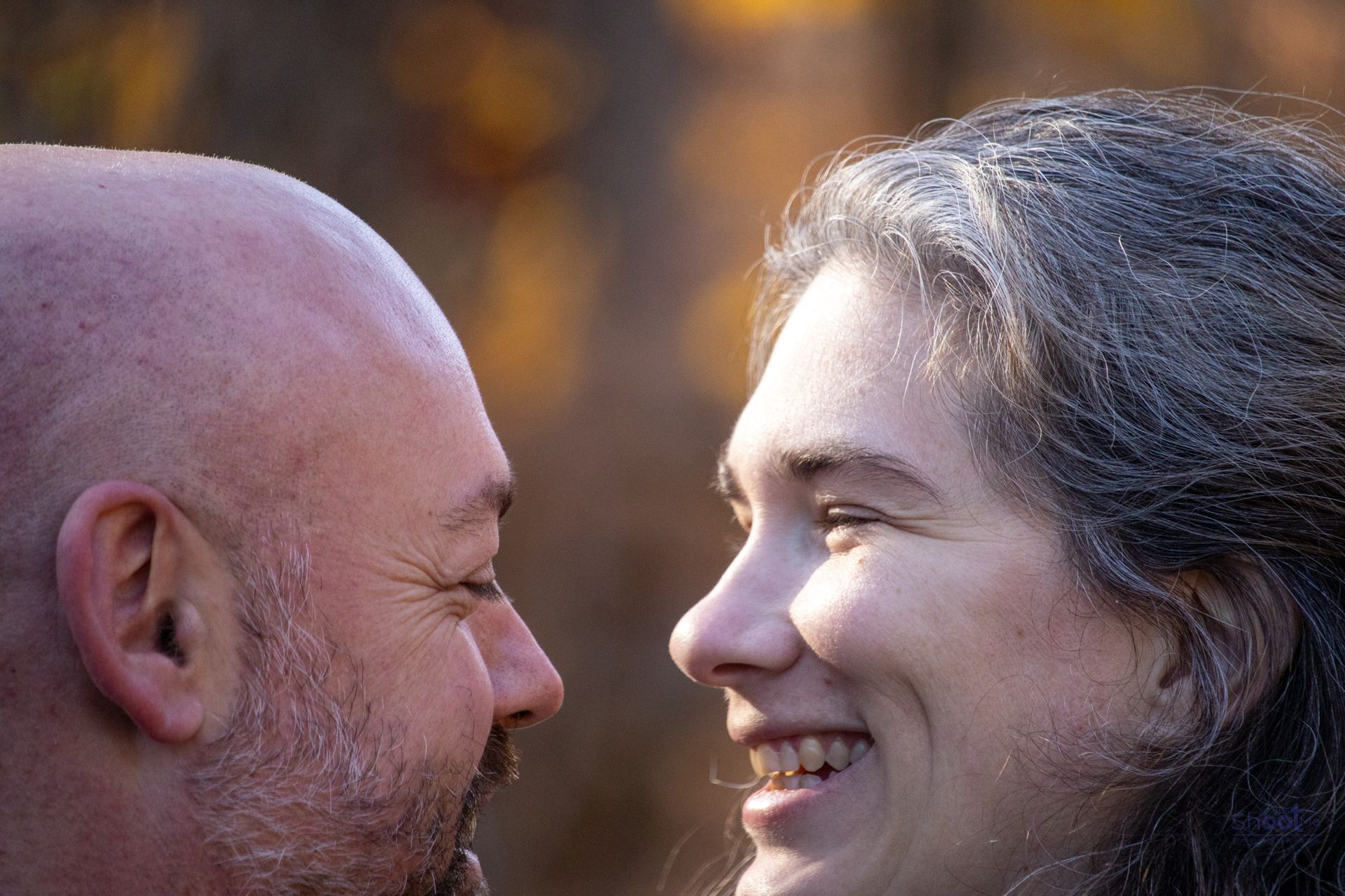 A bald man and a woman with graying hair face each other in profile, smiling softly against a blurred, warm background.