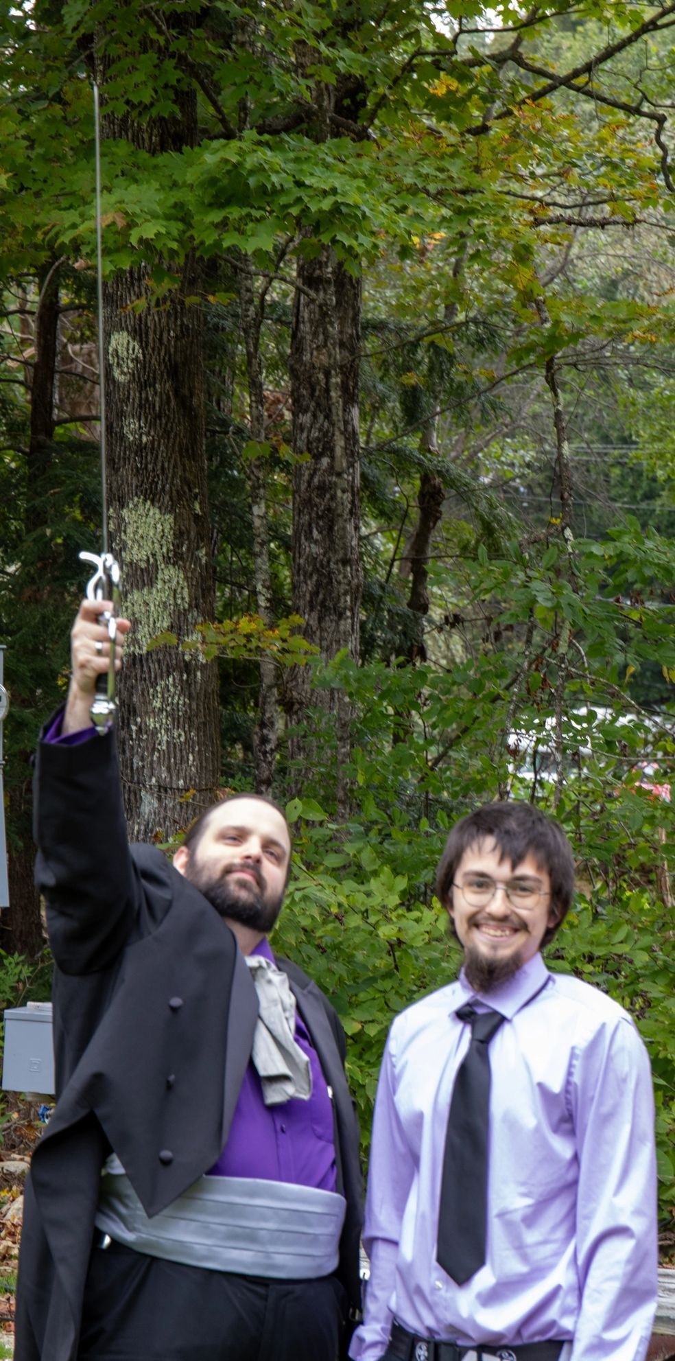 Two men in formal wear stand outdoors; one holds a sword toward the sky, the other smiles beside him.
