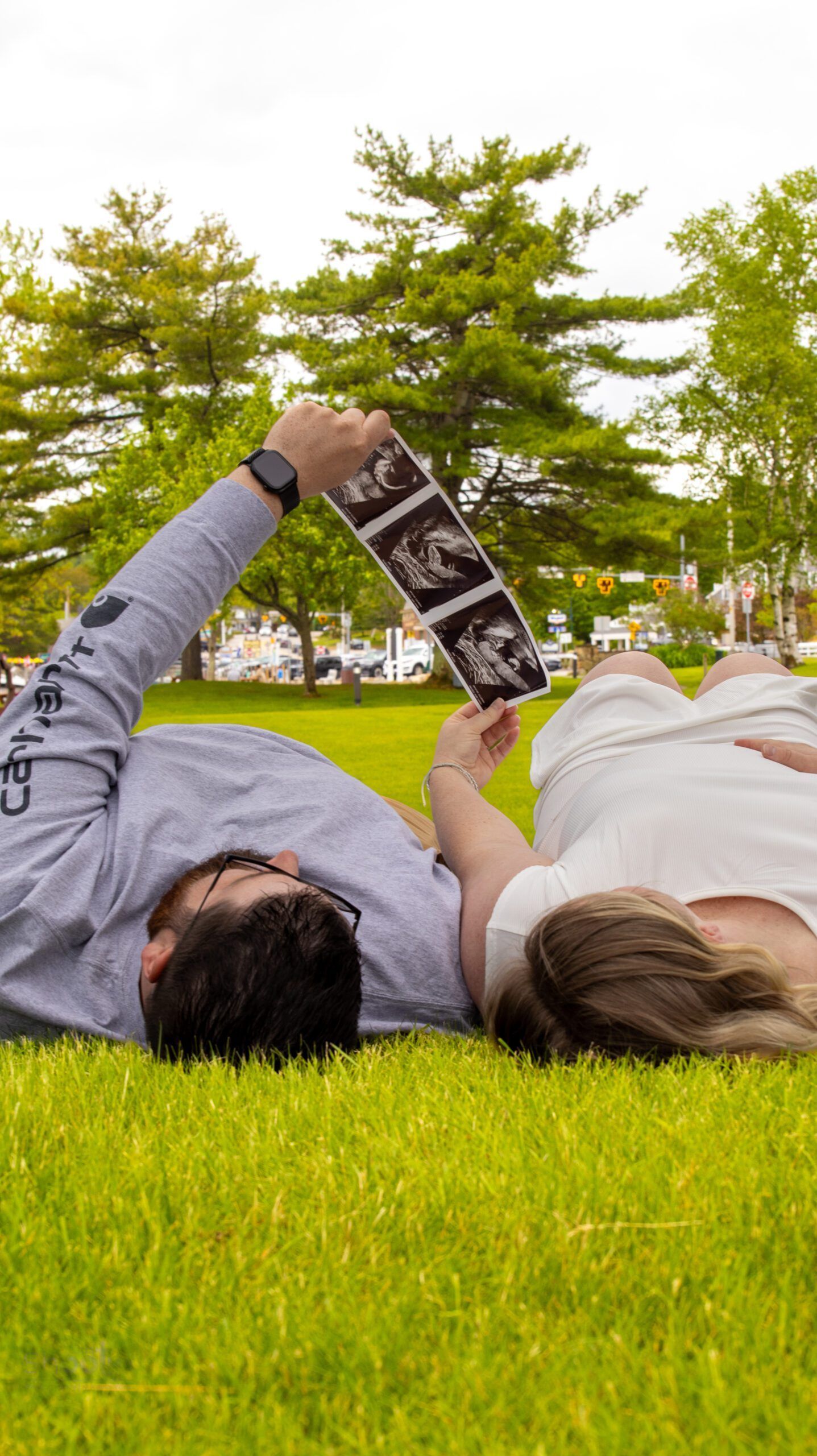 A couple lies in the grass, holding up ultrasound photos towards the sky in an outdoor setting.