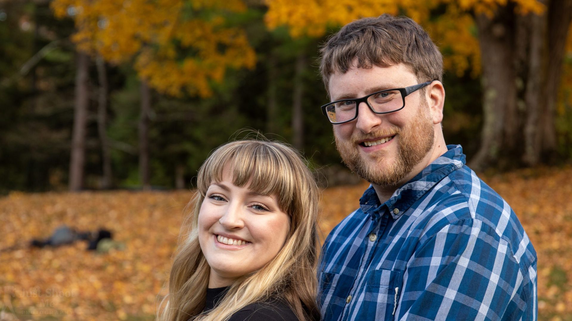 A smiling couple poses for a portrait outdoors in front of a backdrop of autumn leaves.