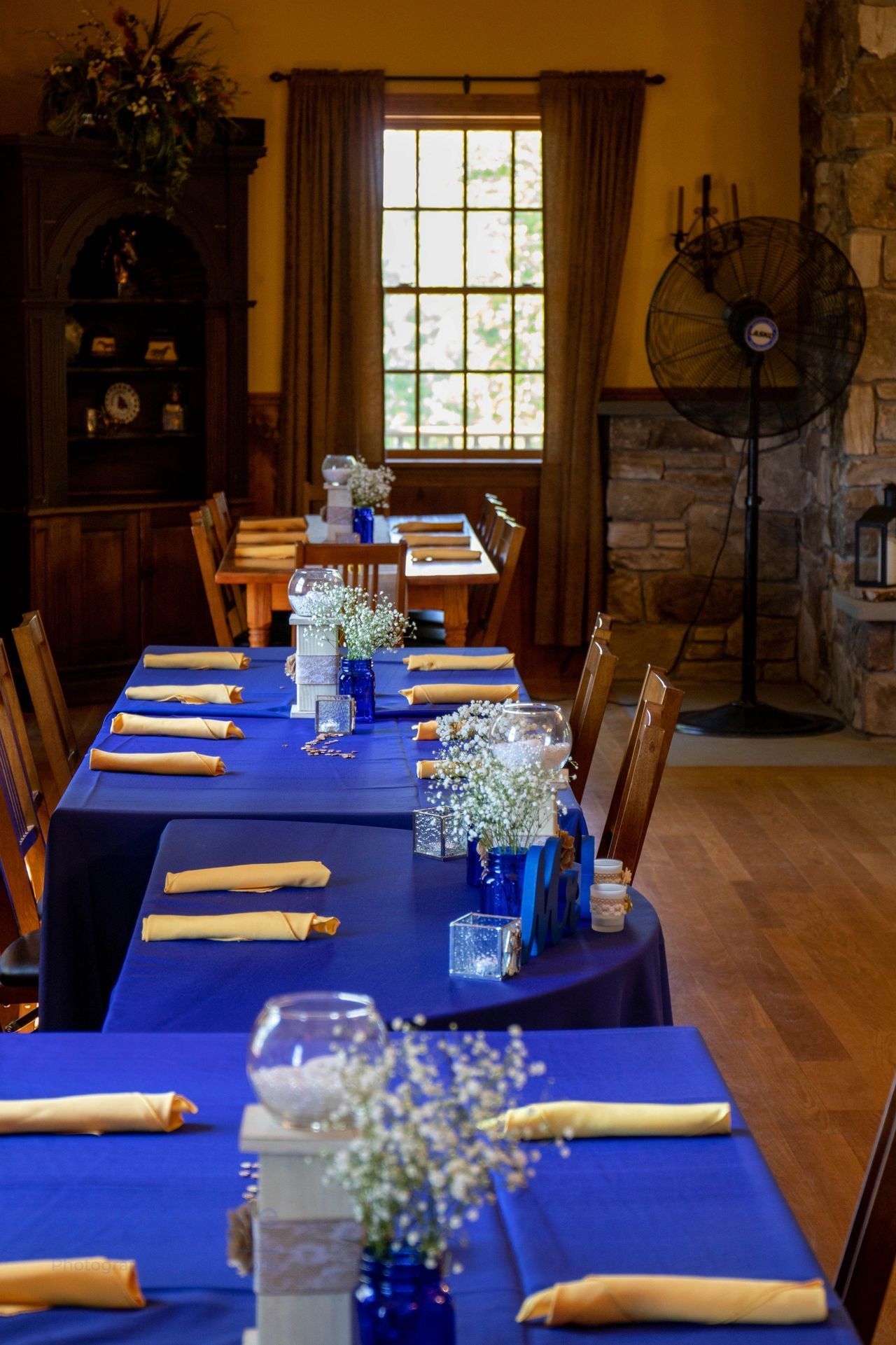 Tables set for a dining event in a room with a large window, stone wall, and dark furniture.