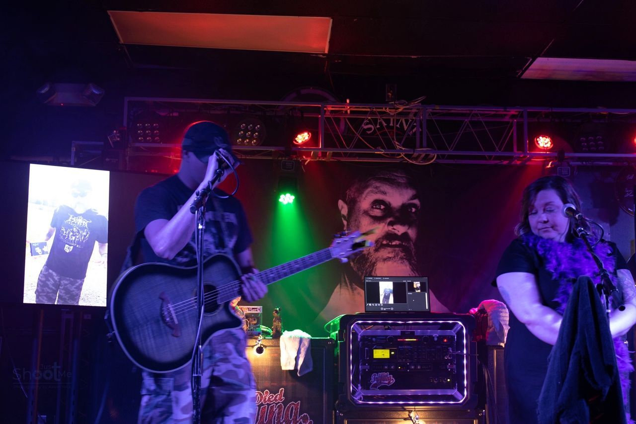 A person playing an acoustic guitar and singing on stage next to another person, with purple and green stage lights.