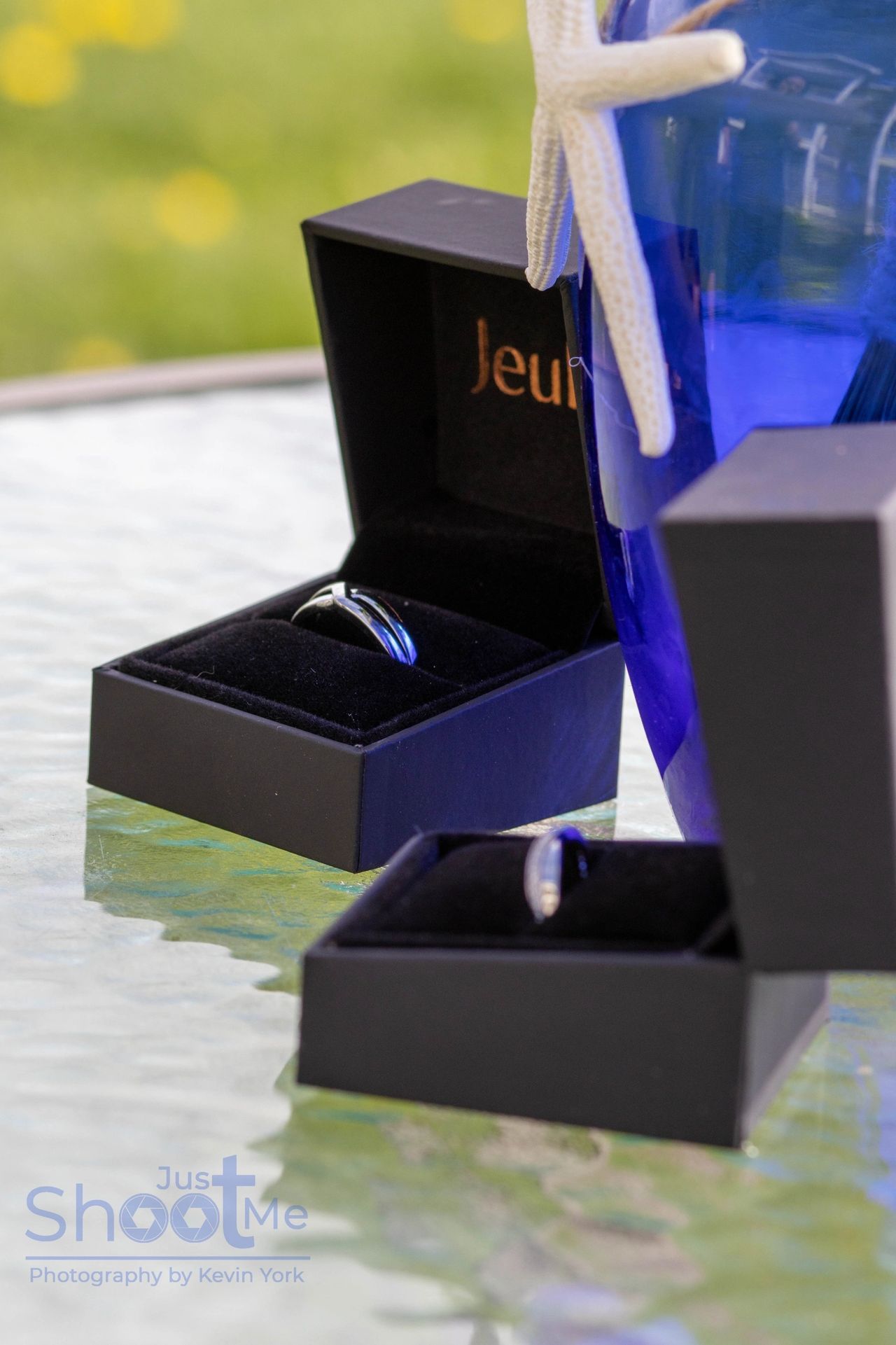 Two rings displayed in open black jewelry boxes, placed next to a blue vase with a white starfish on a glass table.