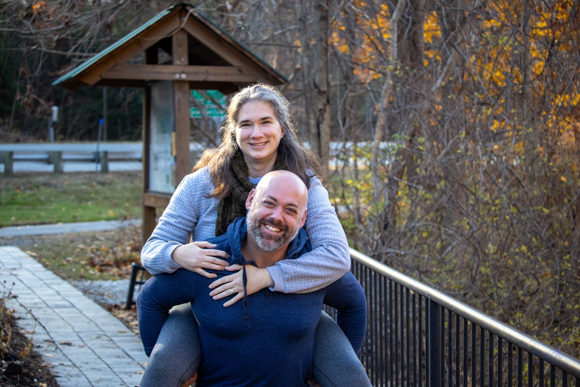 A person carrying another person on their back outdoors, both smiling, in a park-like setting with a wooden structure.