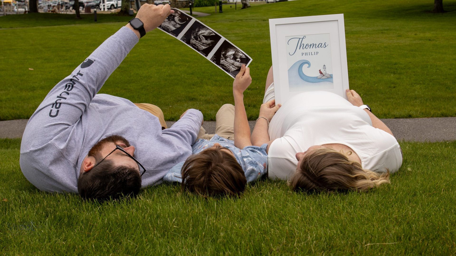 A family lies in the grass, holding an ultrasound scan and a framed announcement featuring a wave and the word 