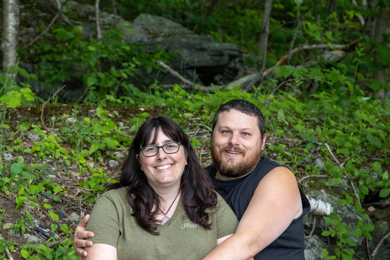 A person with glasses and a person with a beard embrace, smiling in front of a lush, green wooded hillside.