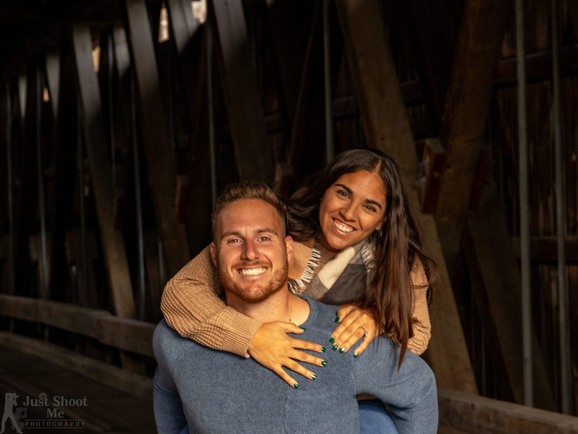 A person giving a piggyback ride to another inside a dark, wooden-structured covered bridge.