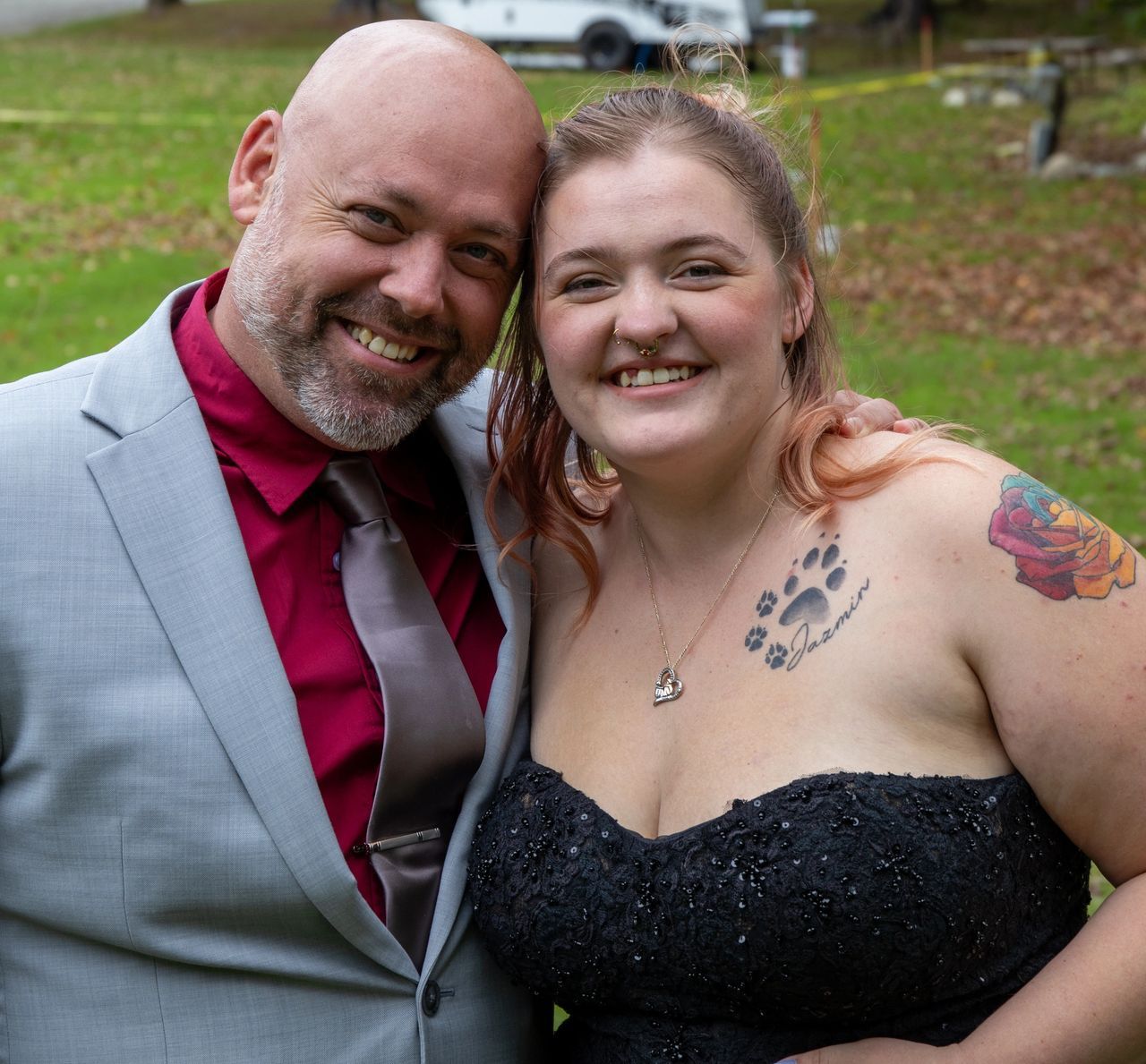 A person in a gray suit and red shirt poses outdoors, head-to-head with a person in a black strapless dress.