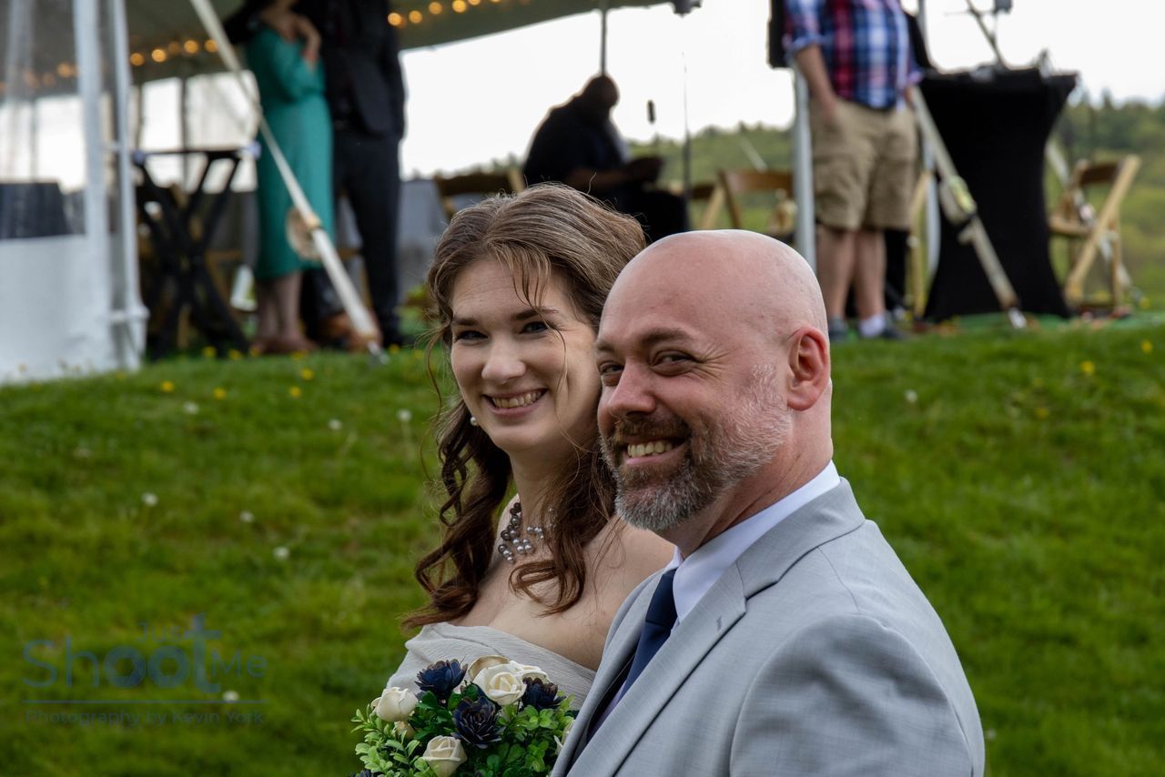 A couple smiles while standing outdoors on a grassy hill during an event held under a white tent.