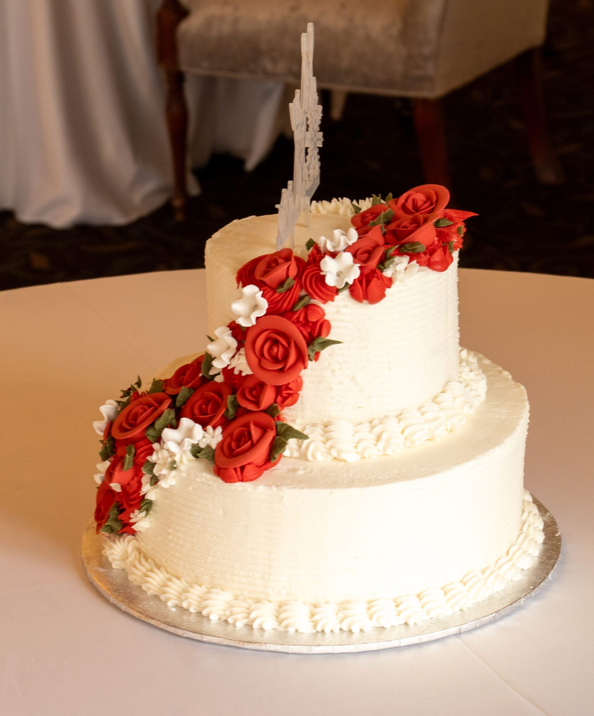 A two-tier white wedding cake decorated with a cascading trail of red roses and white flowers, topped with a castle topper.