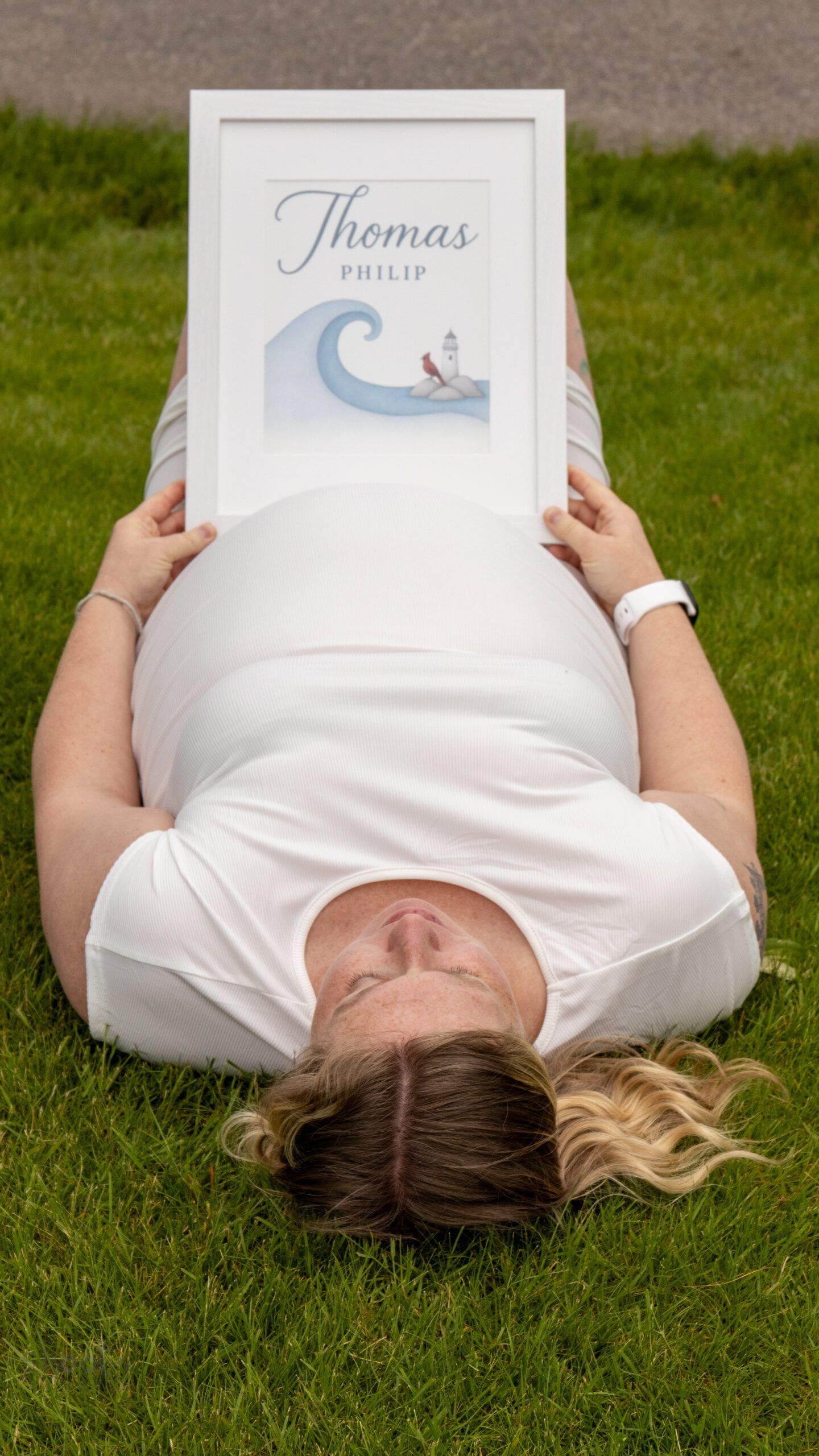A person lying on grass holds a framed print featuring a blue wave, a small boat, and the name 