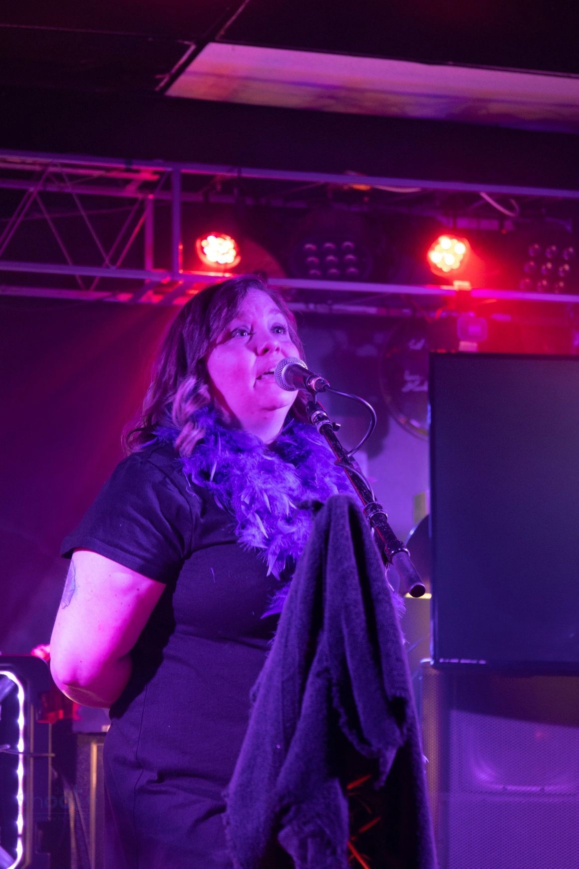 A person in a black shirt and purple feather boa performs into a microphone on a stage with vibrant purple lighting.
