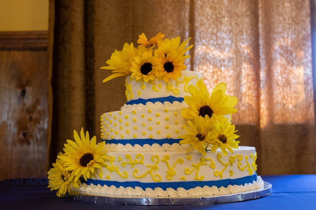 Three-tiered white cake decorated with yellow sunflowers, blue ribbons, and yellow frosting swirls on a blue tablecloth.