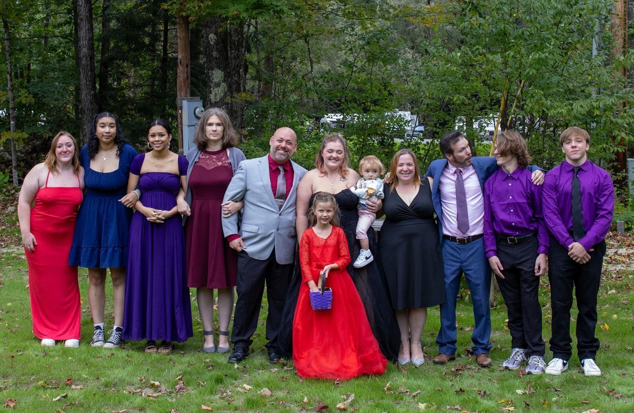A group of 11 people in formal wear pose for a photo in a wooded, grassy outdoor area.