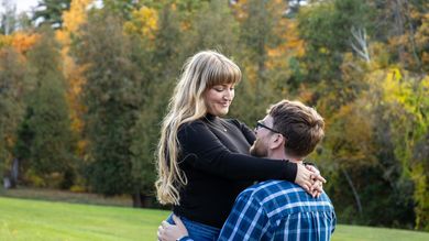 A couple embraces in a grassy field backed by autumn trees, looking at each other with smiles.