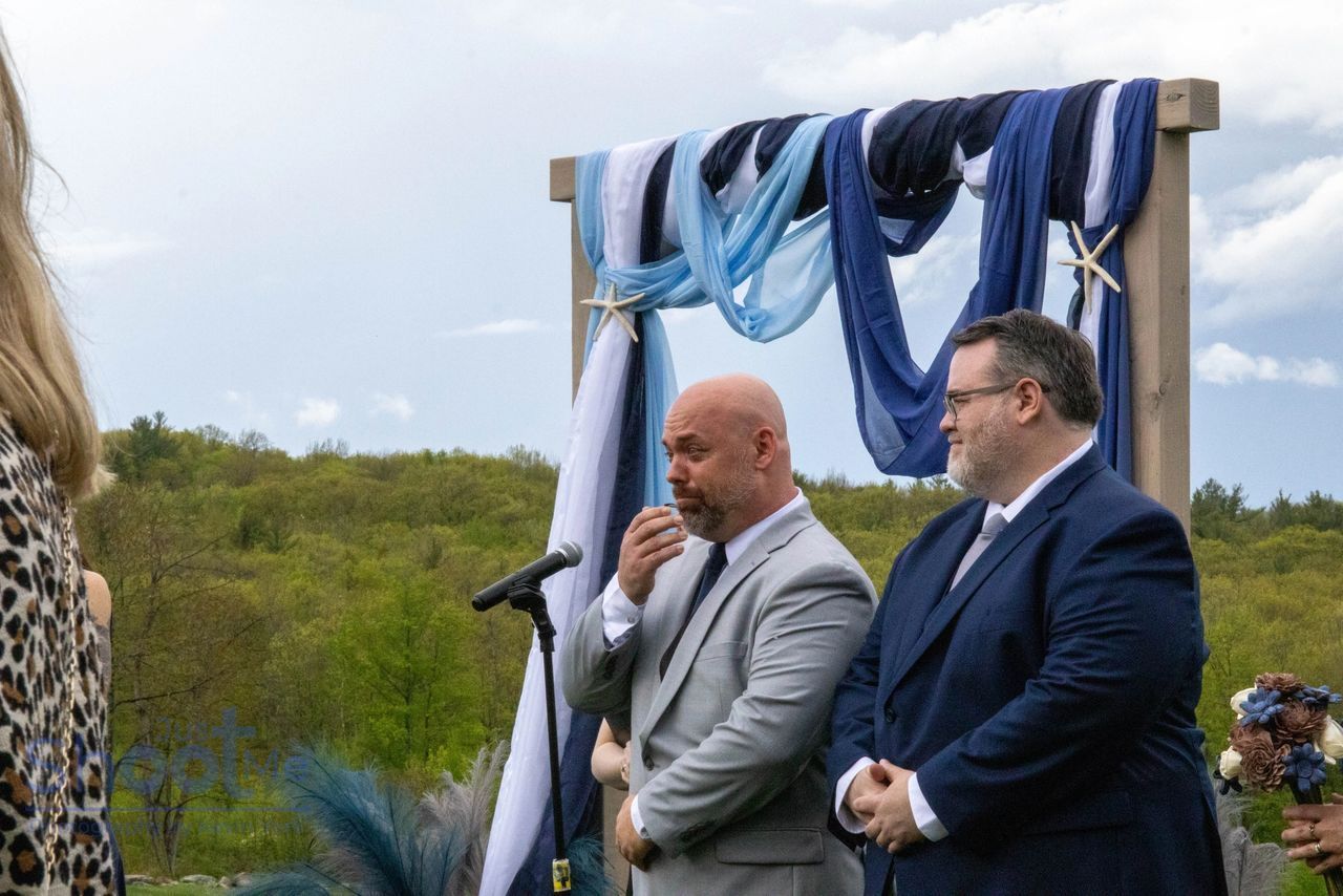 Two people in suits stand before a wedding arbor draped in blue and white fabric, outdoors against a backdrop of trees.