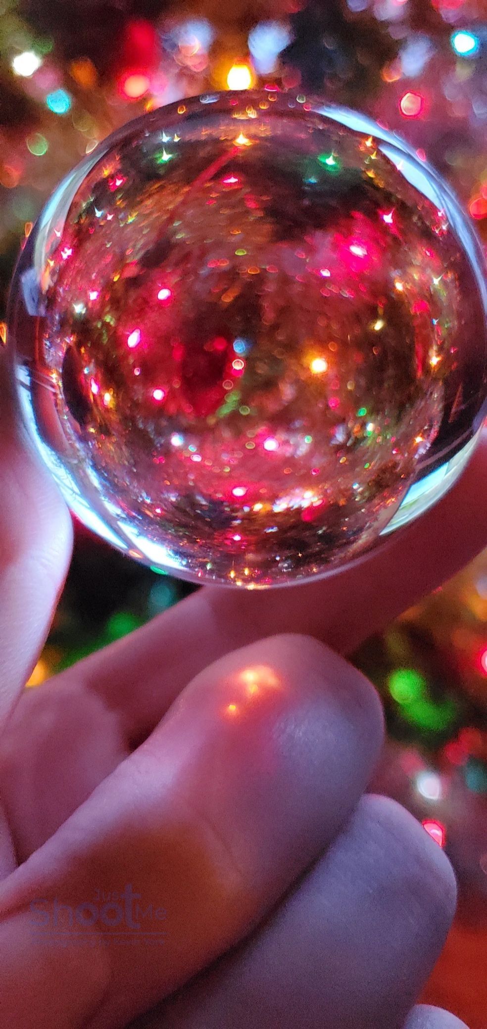 A hand holds a clear glass ornament filled with shimmering, multi-colored light reflections in front of a Christmas tree.
