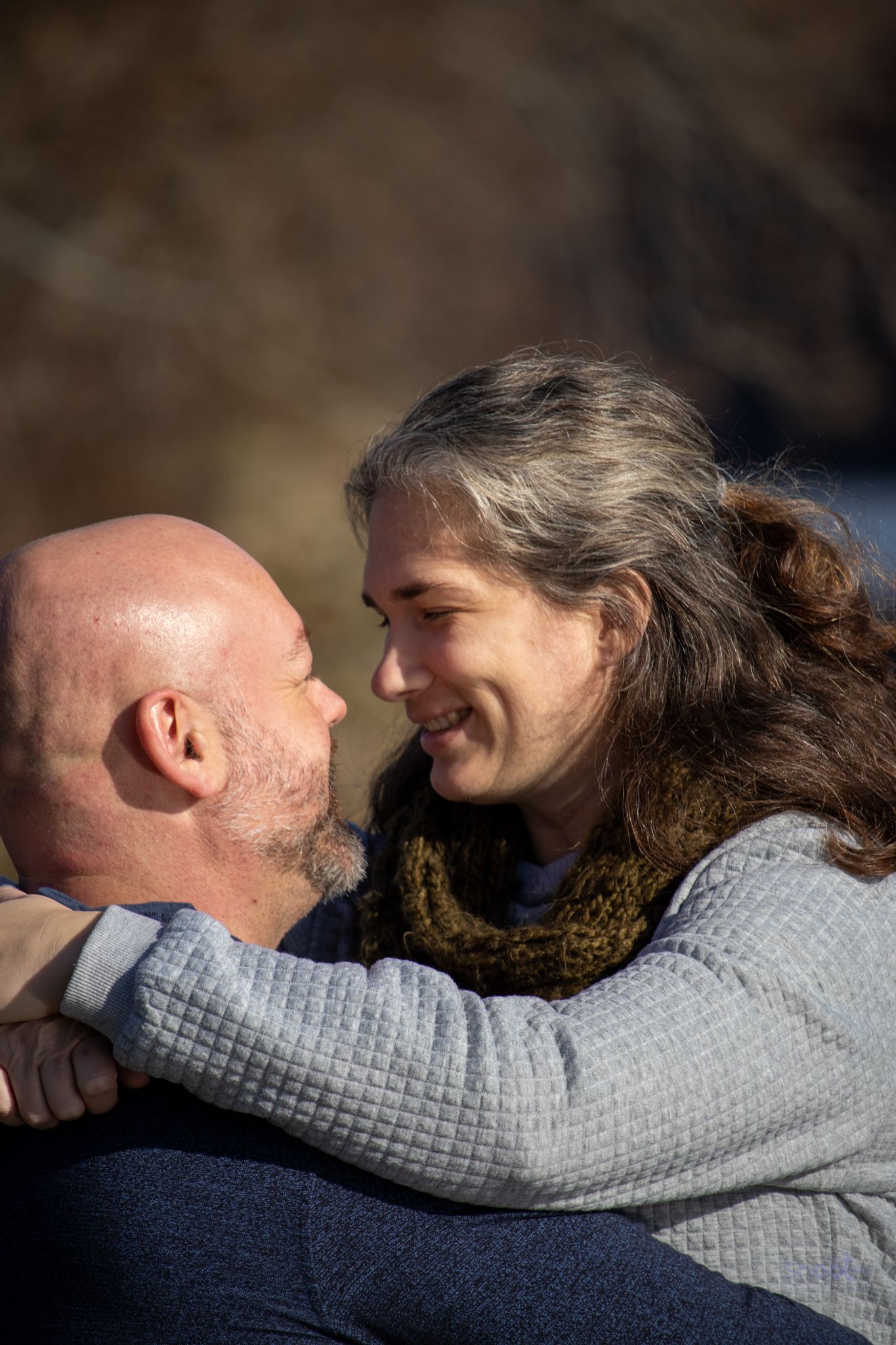 A bald man and a woman with wavy hair embrace and look at each other with soft smiles outdoors.